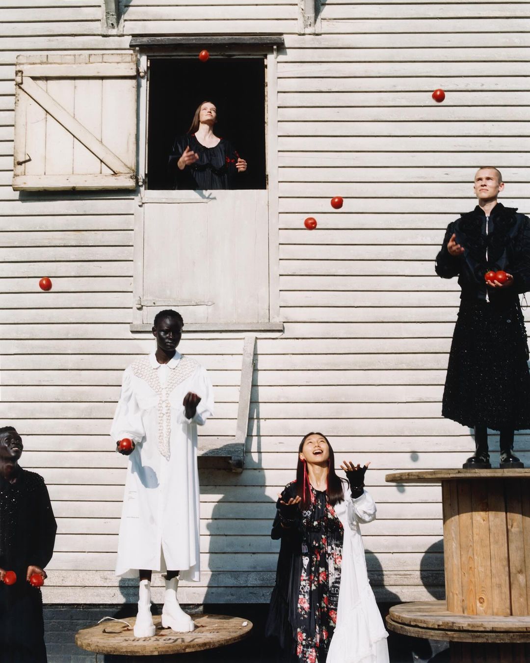 Editorial fashion shot featuring a group of models standing on wooden spools and platforms, juggling red balls in an outdoor setting.