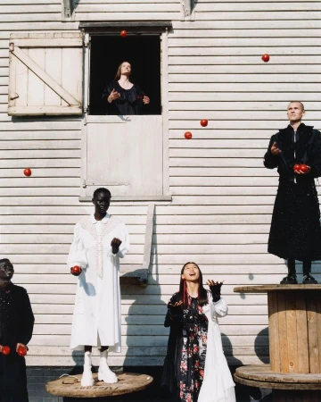 Editorial fashion shot featuring a group of models standing on wooden spools and platforms, juggling red balls in an outdoor setting.