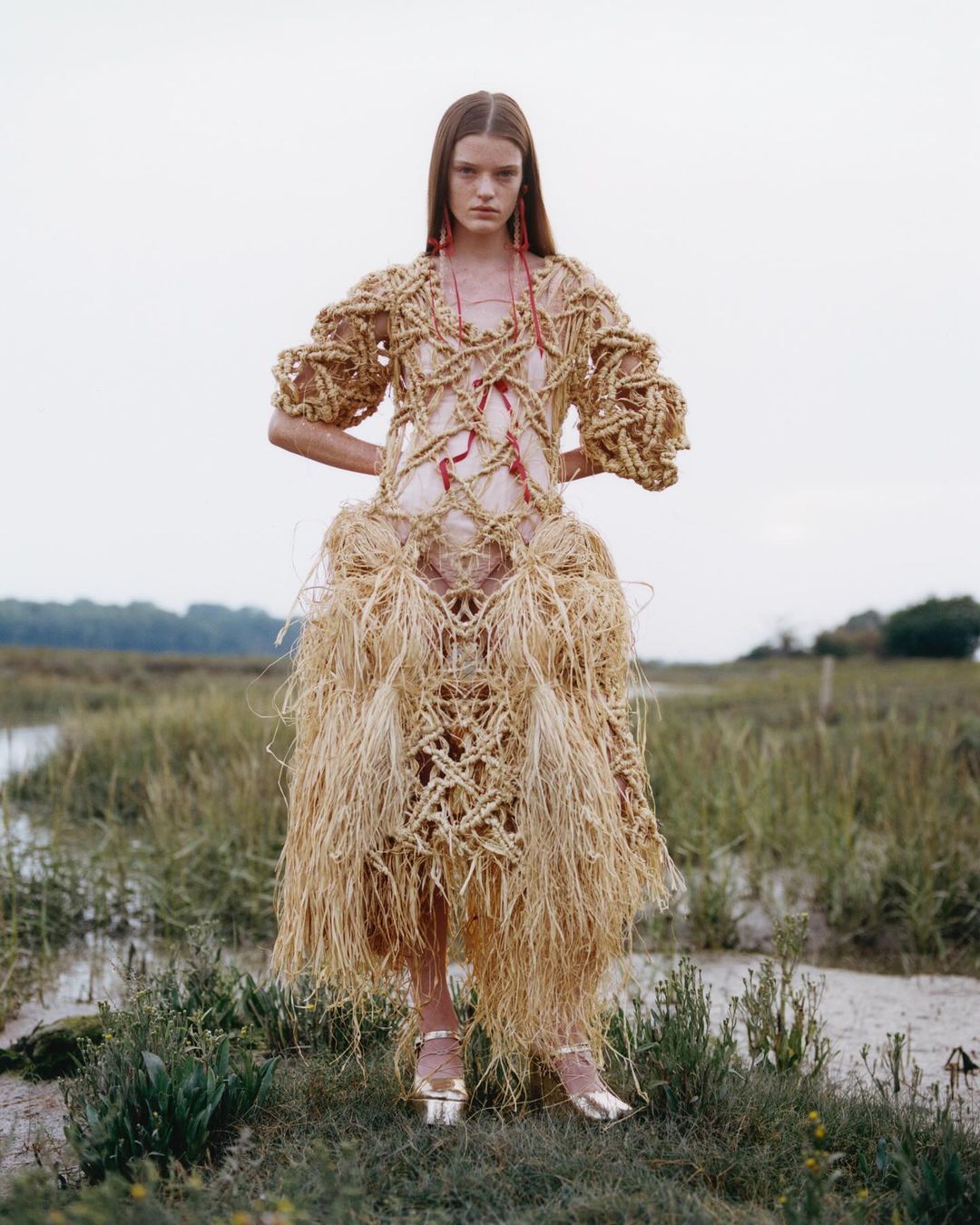 Fashion shot of a woman wearing a woven dress with a grass skirt standing in a grassy field with a body of water in the background.