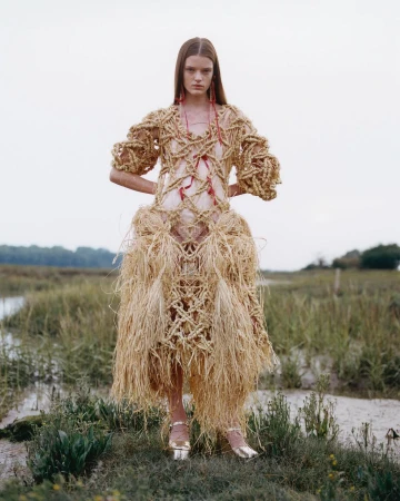 Fashion shot of a woman wearing a woven dress with a grass skirt standing in a grassy field with a body of water in the background.