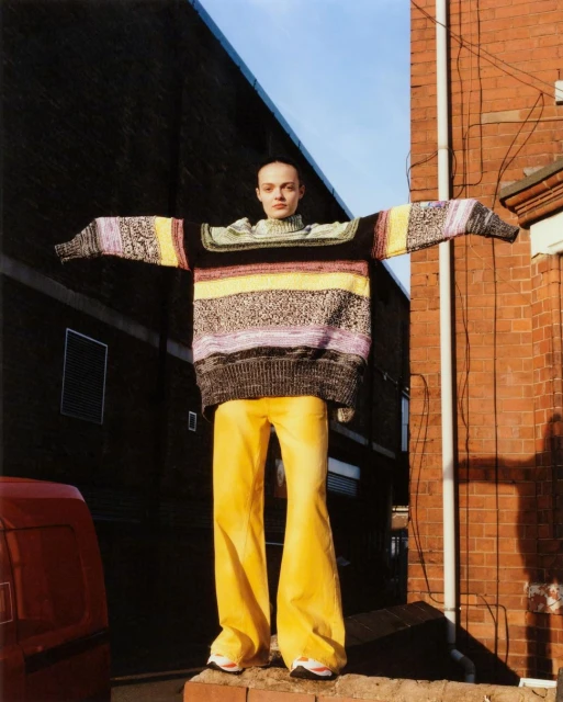 A young woman stands on a low wall in an urban setting, wearing a multicolored sweater and yellow pants, with arms outstretched.