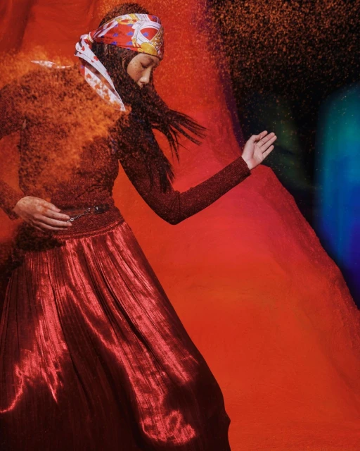 Woman in red metallic dress and patterned headscarf poses with dynamic red powder effect against a gradient red and blue background.