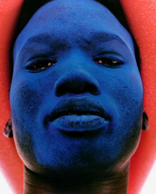Close-up portrait of a human face painted blue, with a soft-focus orange frame around the head, creating a striking, artistic headshot.