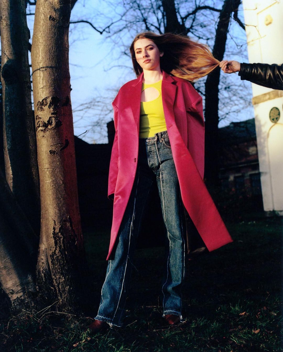 A long-haired woman in jeans and a bright pink coat stands in front of trees and a building with a hand tugging her hair.