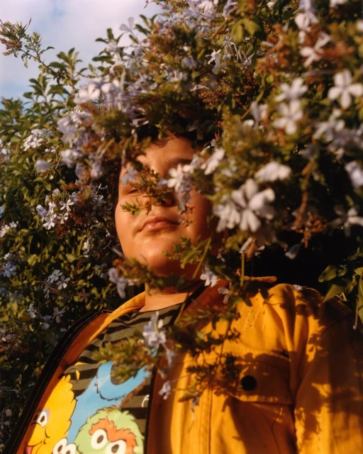 Person surrounded by flowers in outdoor setting, wearing yellow jacket and printed Sesame Street shirt, bathed in soft natural light.