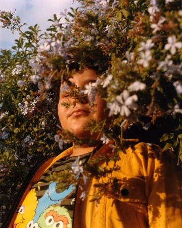 Person surrounded by flowers in outdoor setting, wearing yellow jacket and printed Sesame Street shirt, bathed in soft natural light.