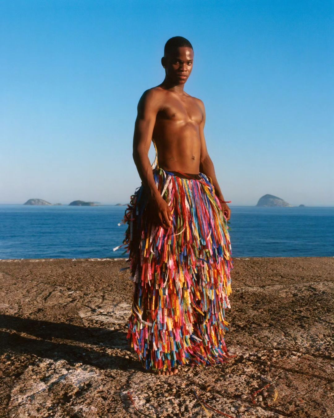 A young Black man stands outdoors in front of the ocean, wearing a decorative skirt made of colorful ribbons, on a sunny day.