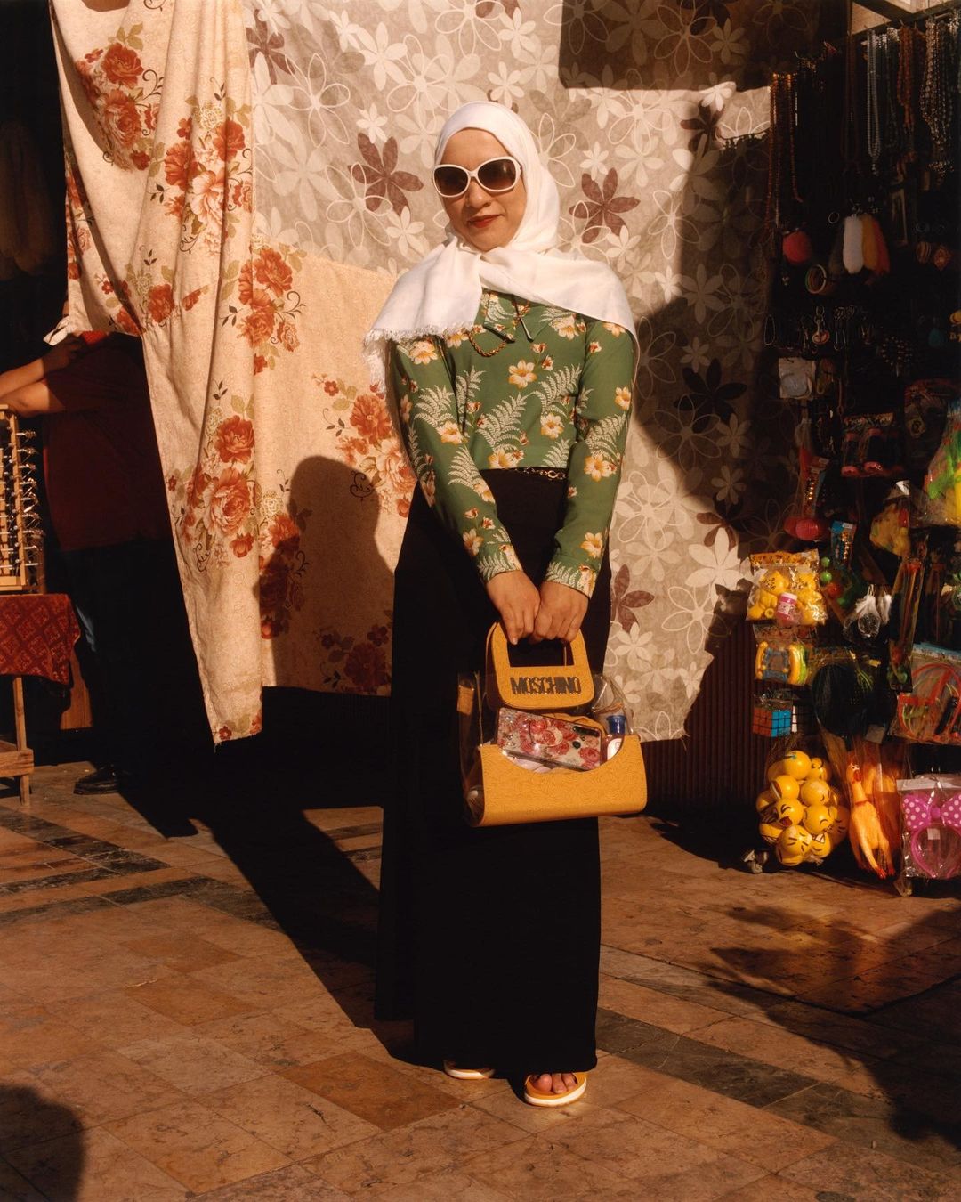 A stylish adult female poses outdoors with a Moschino purse, floral blouse, dark skirt, headscarf, and sunglasses, standing on a stone surface.