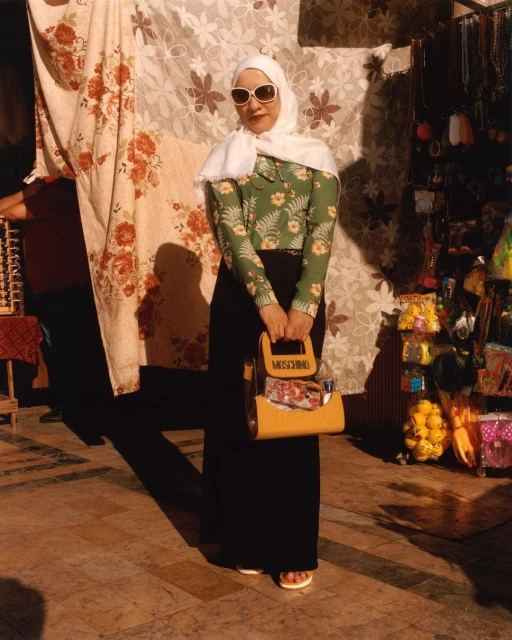 A stylish adult female poses outdoors with a Moschino purse, floral blouse, dark skirt, headscarf, and sunglasses, standing on a stone surface.