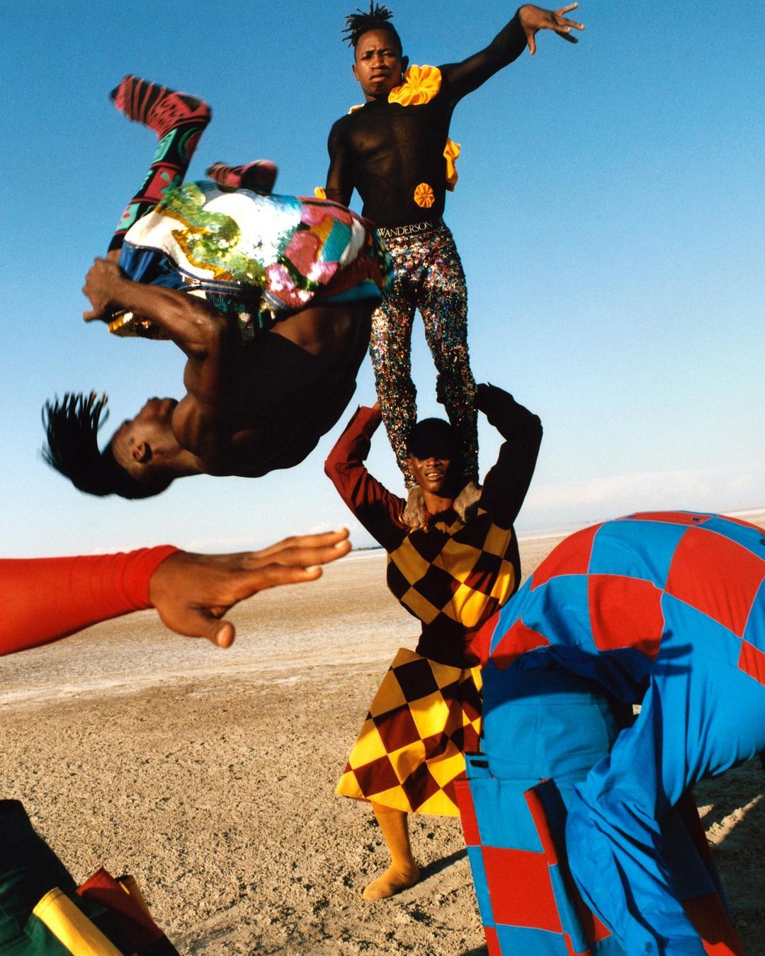 Group of male dancers in brightly colored costumes perform acrobatic poses on a flat, sandy landscape under a clear, light blue sky.