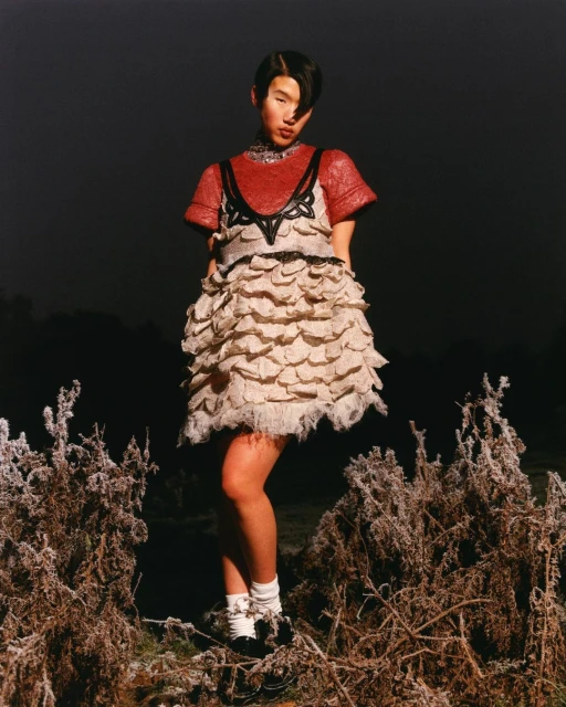 A full shot shows a woman in an avant-garde dress, standing outdoors amidst frost-covered plants under an overcast sky, styled for fashion.