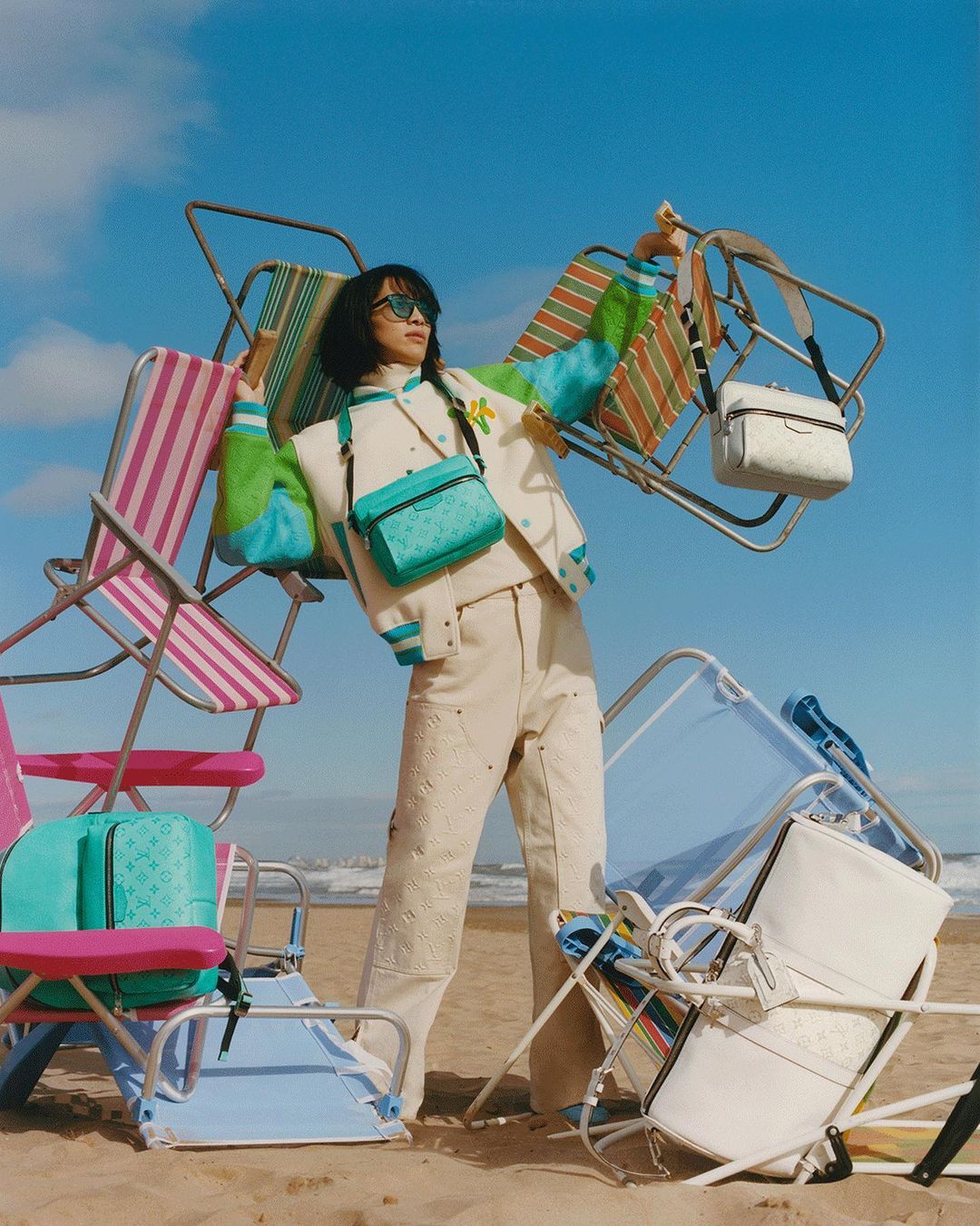 A woman wearing a varsity jacket, trousers, and sunglasses stands on a beach surrounded by overturned beach chairs and handbags in a fashion shoot.