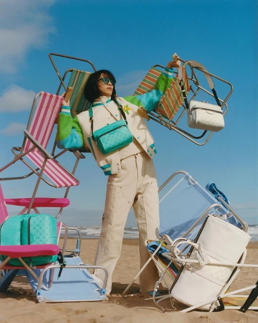 A woman wearing a varsity jacket, trousers, and sunglasses stands on a beach surrounded by overturned beach chairs and handbags in a fashion shoot.