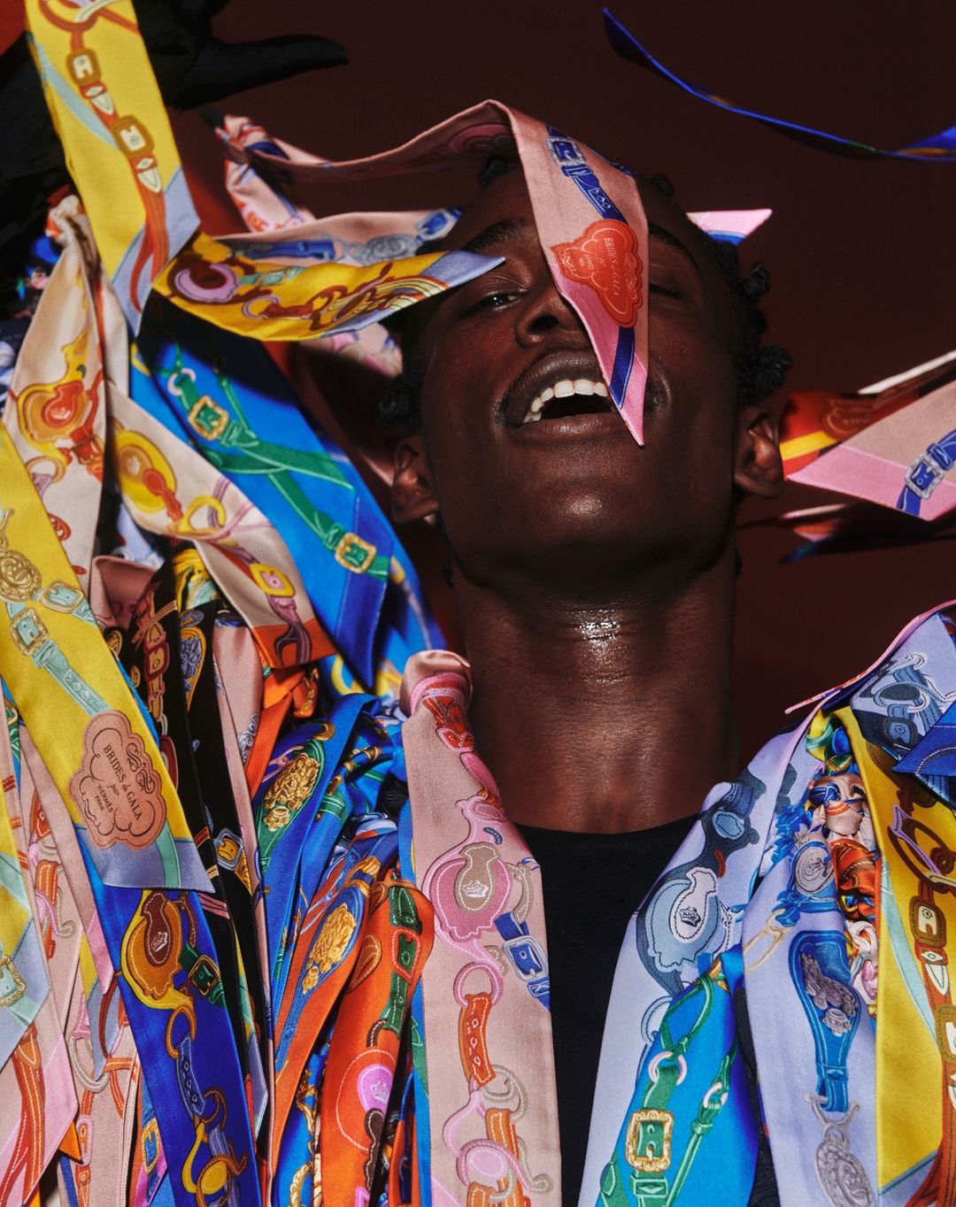 Close-up studio portrait of a Black male model with a collection of patterned scarves draped around him and overhead, against a brown background.