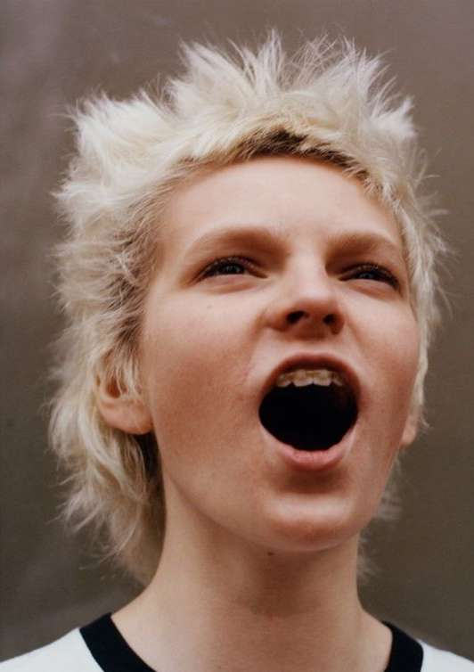 Low-angle portrait of a young adult with blond spiky hair and braces, mouth open, against a neutral gray background in a studio.