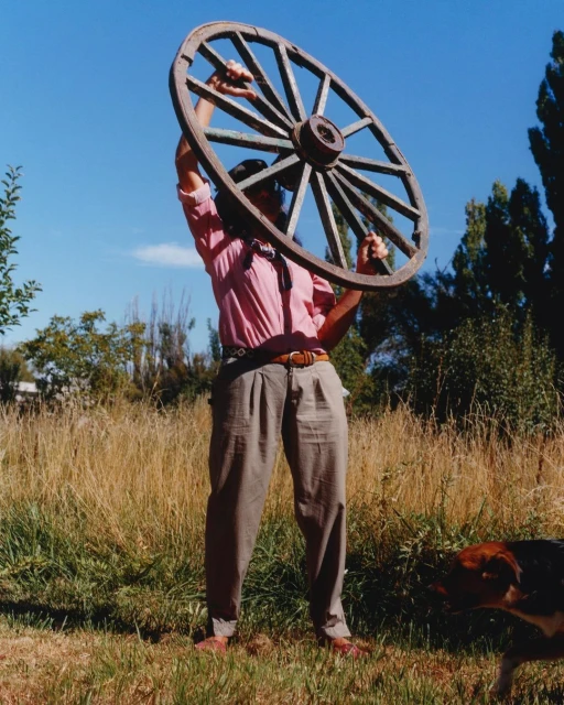 Person in rural setting holds antique wooden wagon wheel overhead against a blue sky backdrop on a sunny day, dog in foreground.