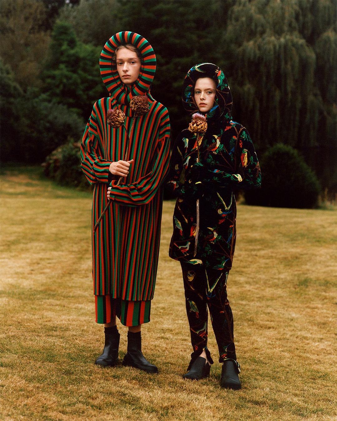 Two people in hooded striped and patterned robes stand in a grassy field with trees, holding thistle flowers, for a fashion editorial.
