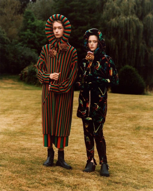 Two people in hooded striped and patterned robes stand in a grassy field with trees, holding thistle flowers, for a fashion editorial.