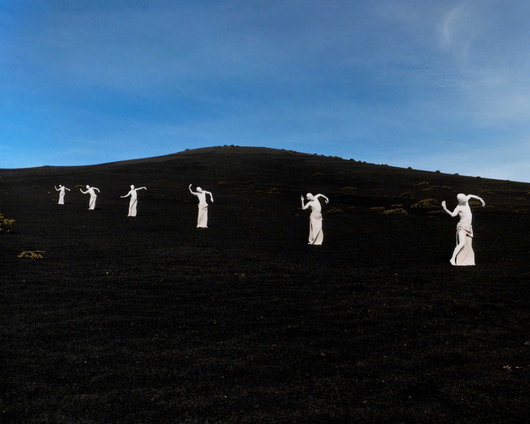 Seven people in white robes pose on a black slope beneath a blue sky, creating an artistic landscape scene with a sense of mystery.