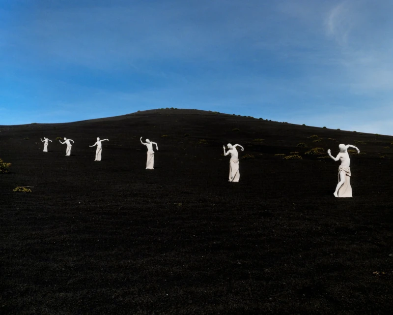 Seven people in white robes pose on a black slope beneath a blue sky, creating an artistic landscape scene with a sense of mystery.