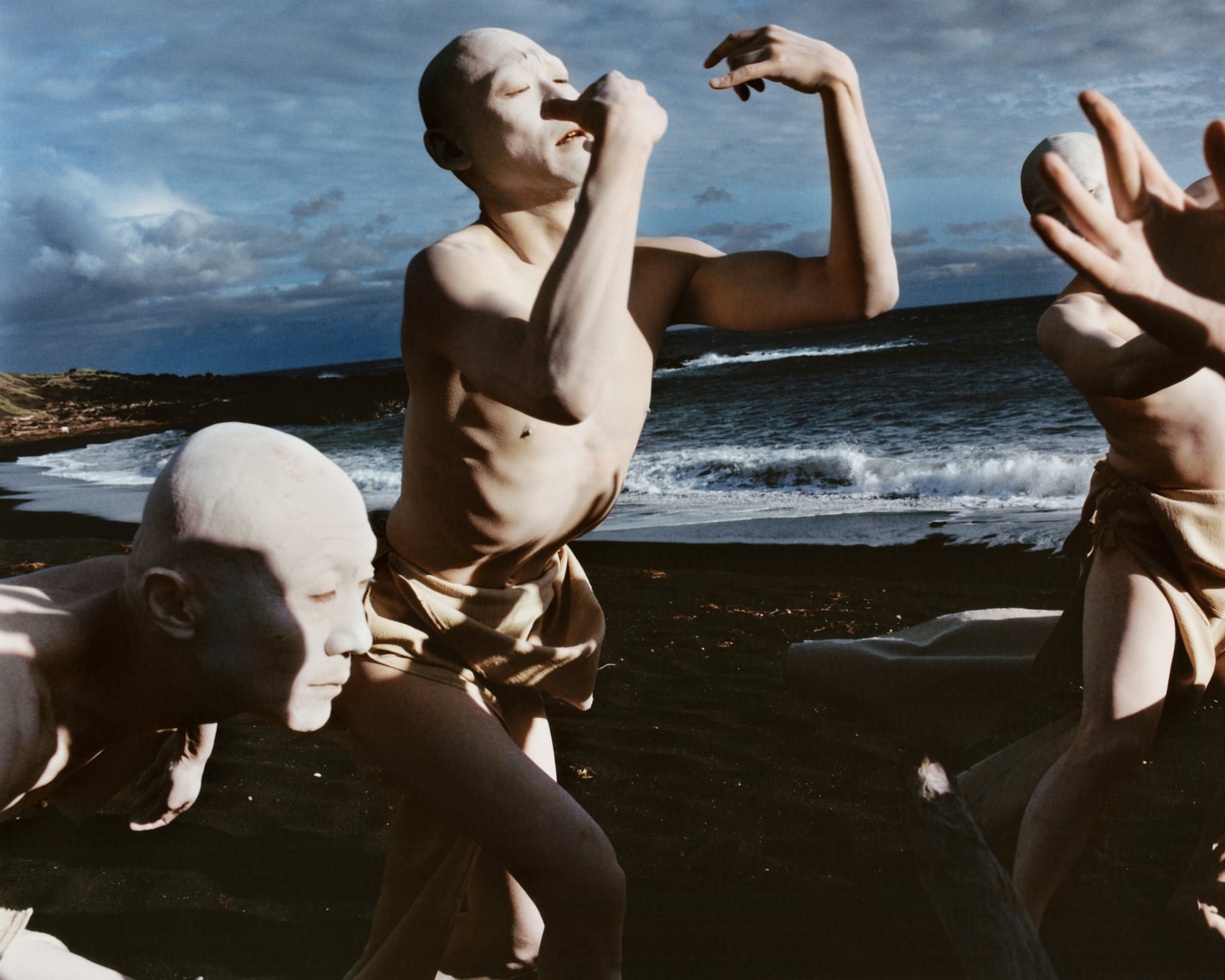 Three figures covered in pale clay pose on a dark sand beach with an ocean backdrop, evoking a primal, artistic aesthetic.