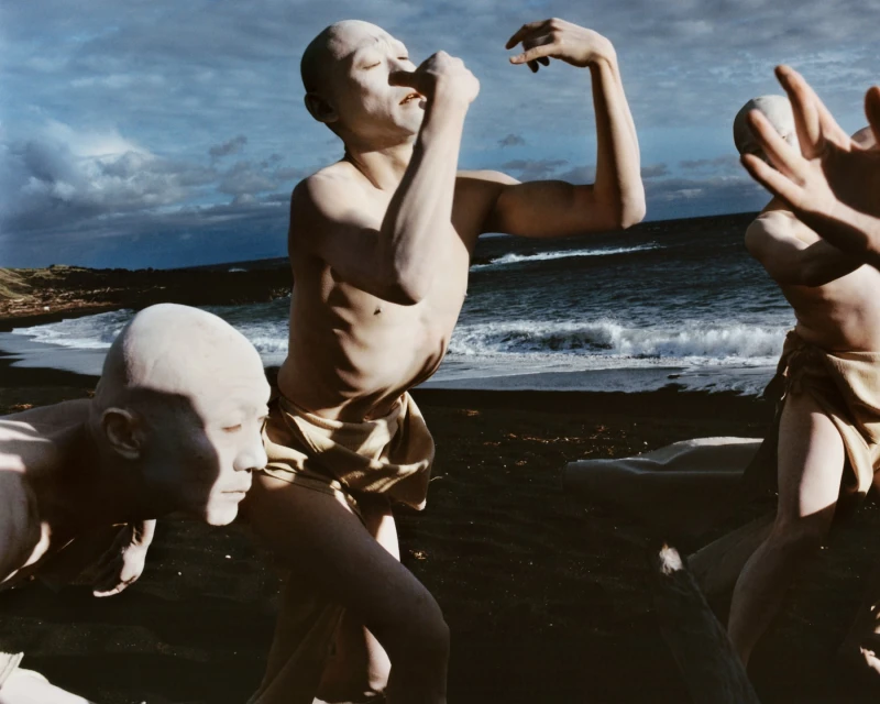 Three figures covered in pale clay pose on a dark sand beach with an ocean backdrop, evoking a primal, artistic aesthetic.
