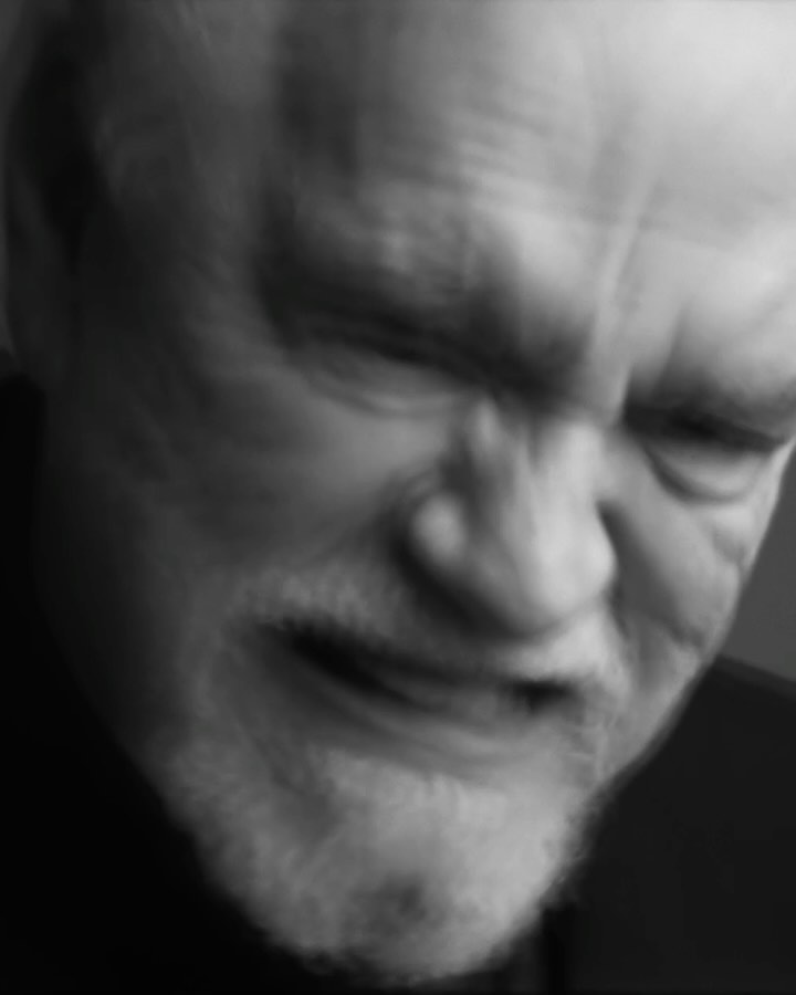Close-up black and white portrait of an adult man with a white beard and dark clothing against a dark, blurred background.