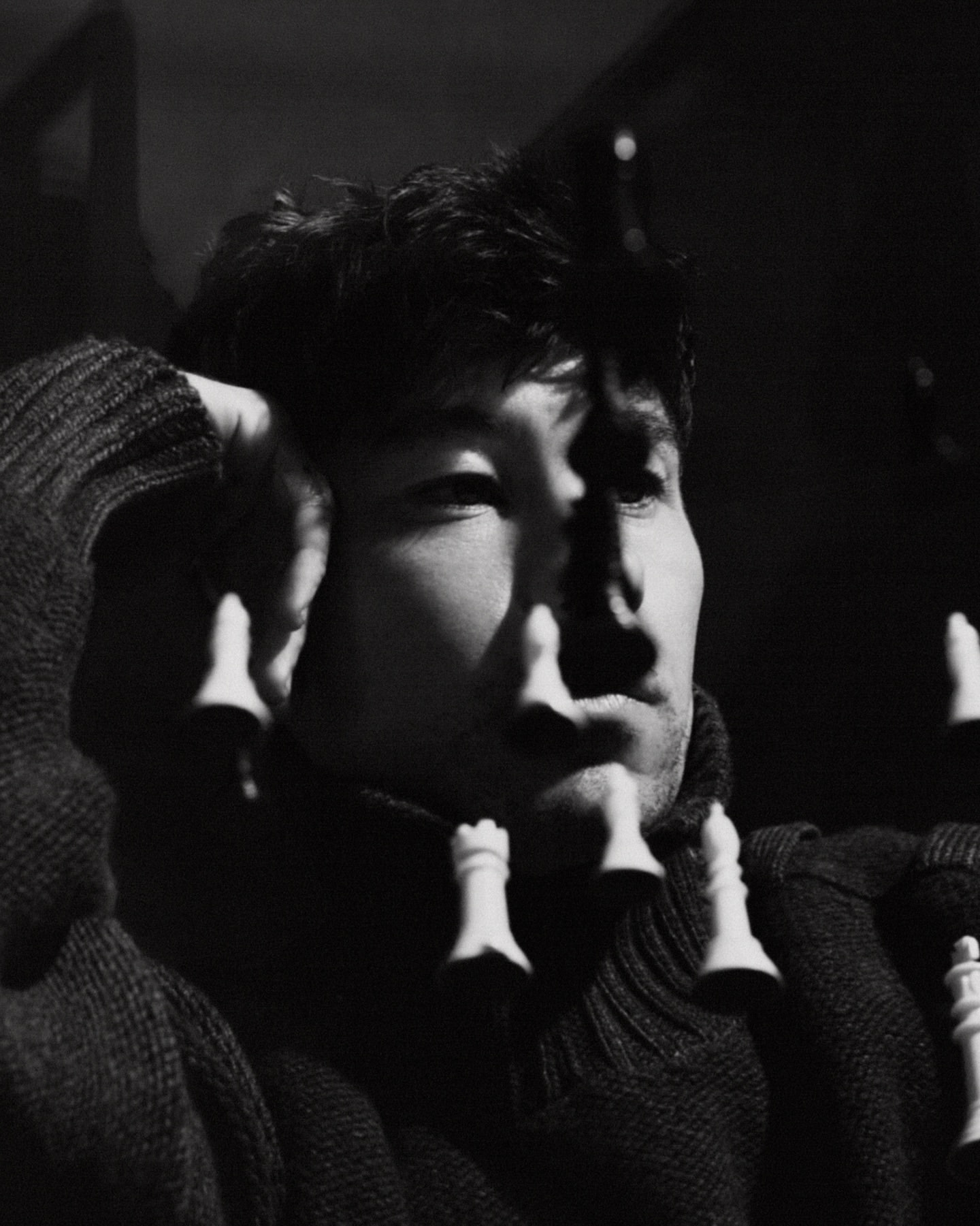 Monochromatic portrait of a man in a sweater with chess pieces suspended in front of his face, showcasing depth and contrast.