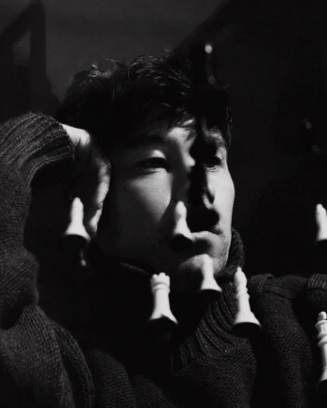 Monochromatic portrait of a man in a sweater with chess pieces suspended in front of his face, showcasing depth and contrast.