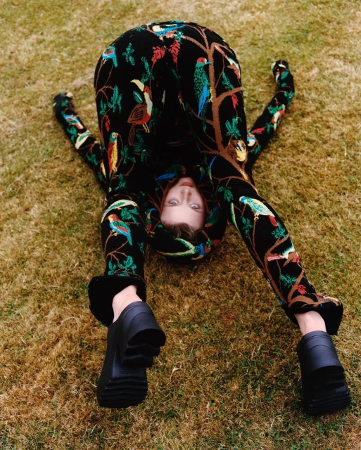 An adult woman in a bird-patterned jumpsuit is laying upside down on grass, wearing black platform shoes in an outdoor fashion pose.