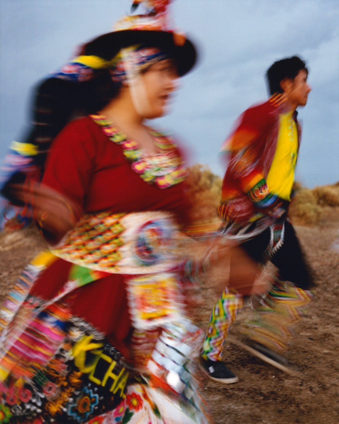 Two people in colorful traditional clothing walk on a dirt path, blurred motion, outdoor setting with overcast sky in the background.