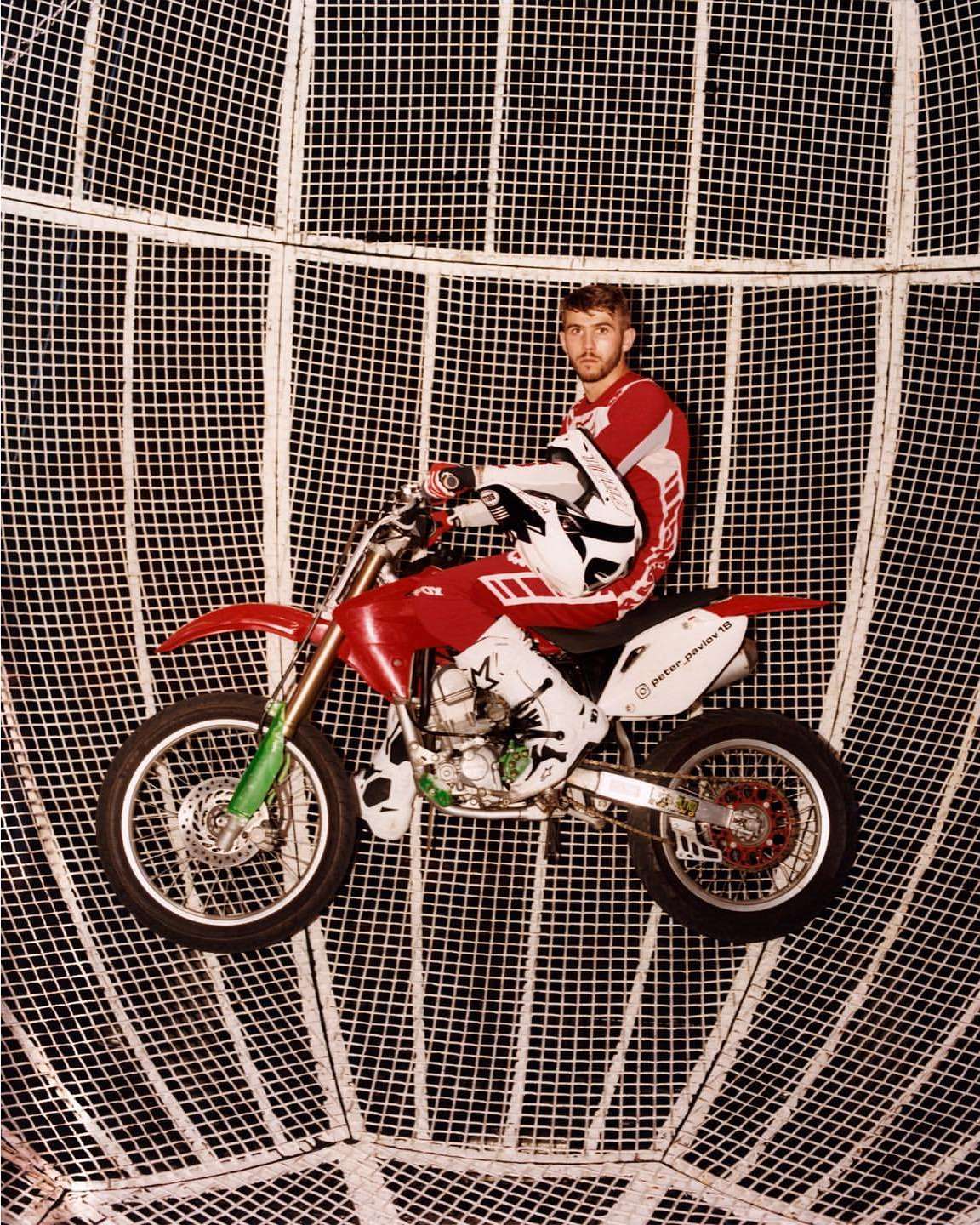 Motocross rider on a red dirt bike performs inside a mesh sphere, wearing red and white gear and a helmet, showcasing action and skill.