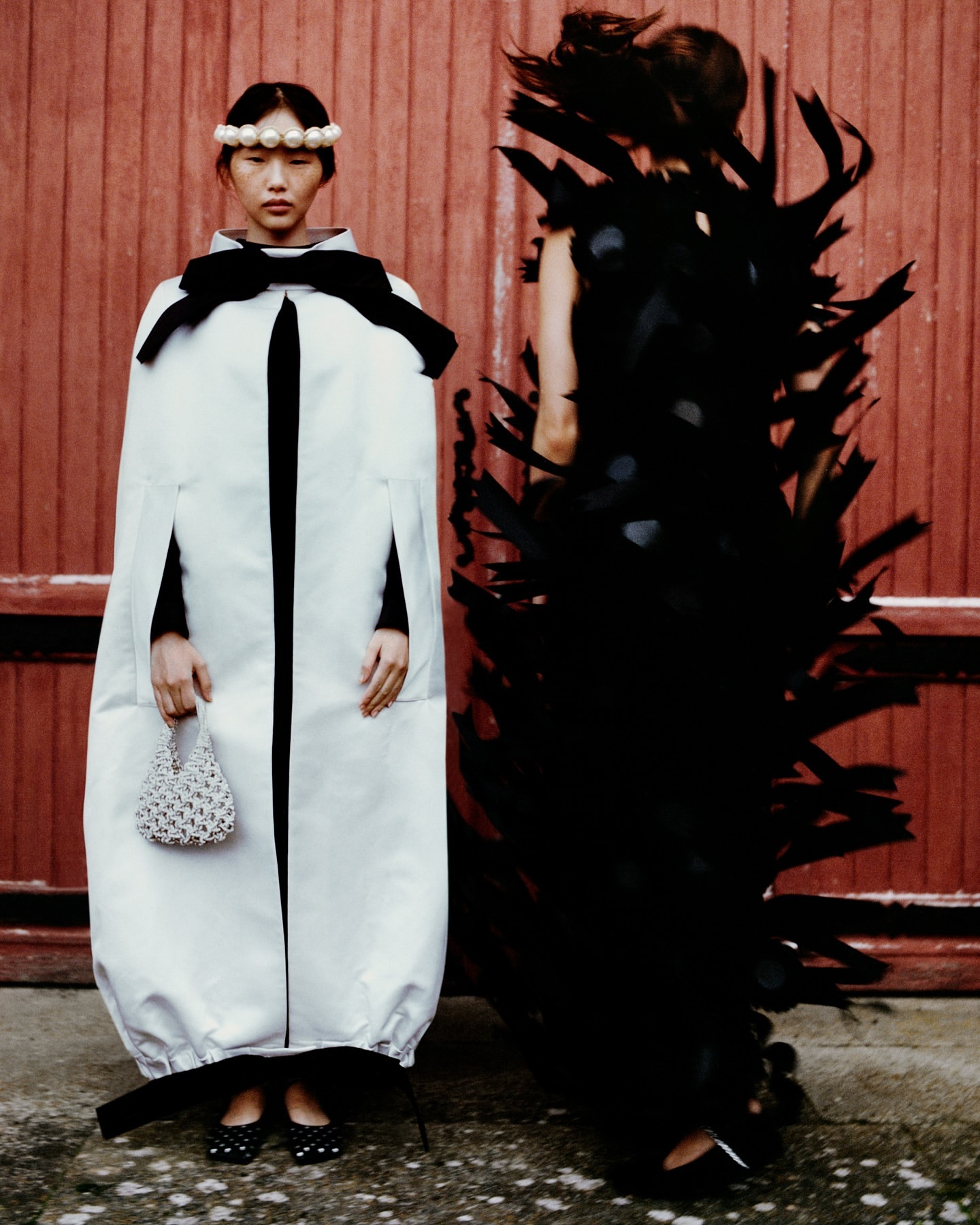 Two women model avant-garde fashion against a red wall. One in white with pearl headband; the other in black feathers, outdoors.