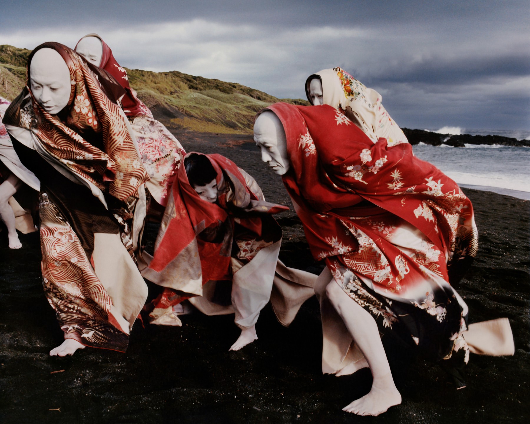 Group of figures with white-painted faces and patterned kimonos on a black sand beach near the ocean and a grassy hill under a cloudy sky.
