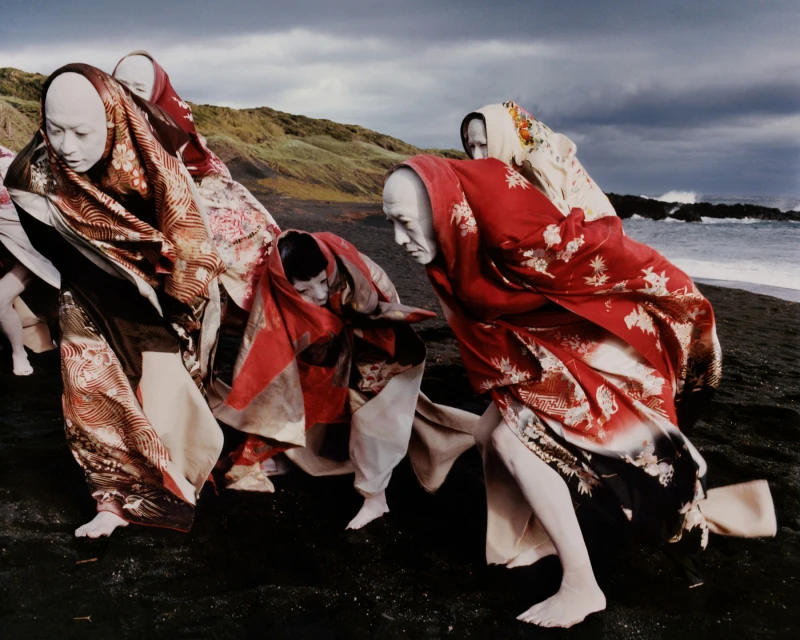 Group of figures with white-painted faces and patterned kimonos on a black sand beach near the ocean and a grassy hill under a cloudy sky.