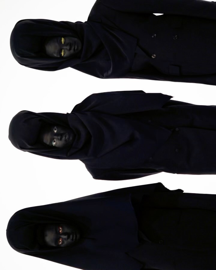 Three hooded figures with darkened skin and striking eyes are stacked vertically against a white background in a studio shot.