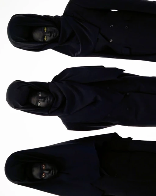 Three hooded figures with darkened skin and striking eyes are stacked vertically against a white background in a studio shot.