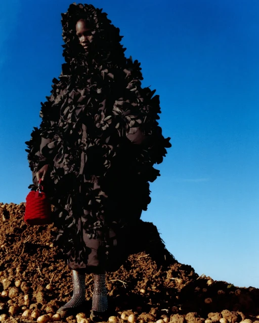 Fashion shot of a woman in a floral hooded coat holding a small red bucket bag standing on a mound of potatoes against a blue sky.