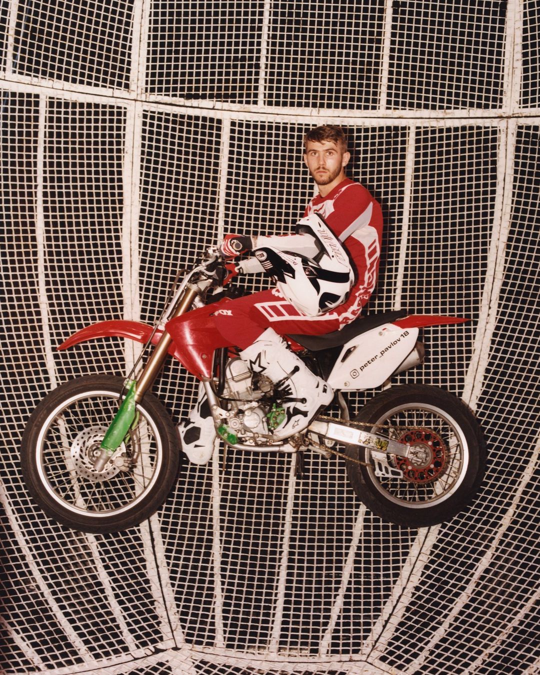 Male motorcyclist in red and white gear on a red and silver dirtbike, inside of a metal cage sphere, posing for a studio shot.