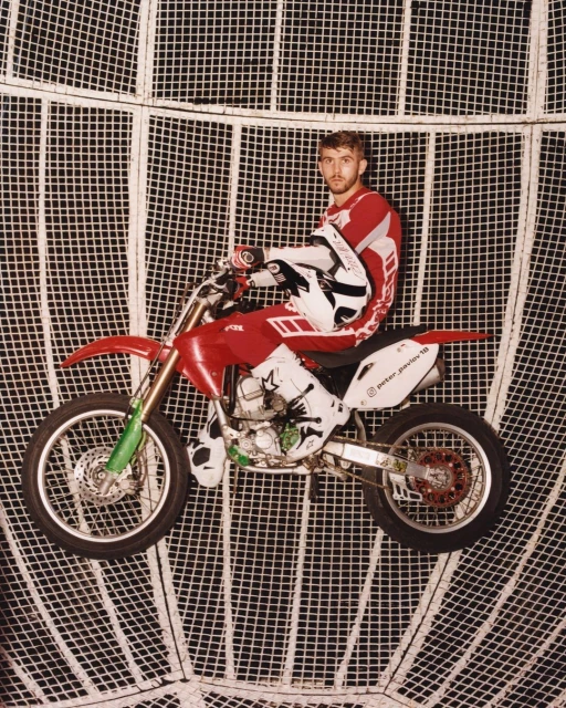 Male motorcyclist in red and white gear on a red and silver dirtbike, inside of a metal cage sphere, posing for a studio shot.