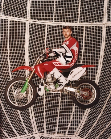 Male motorcyclist in red and white gear on a red and silver dirtbike, inside of a metal cage sphere, posing for a studio shot.