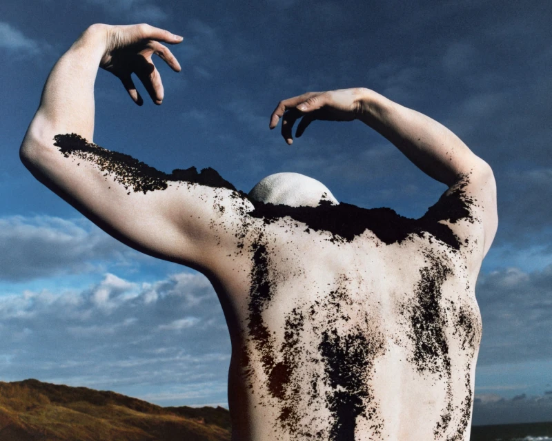 Artistic nude photo of a male figure from the back, covered in dark sand or earth, set against a blue sky with light clouds and a distant hillside.