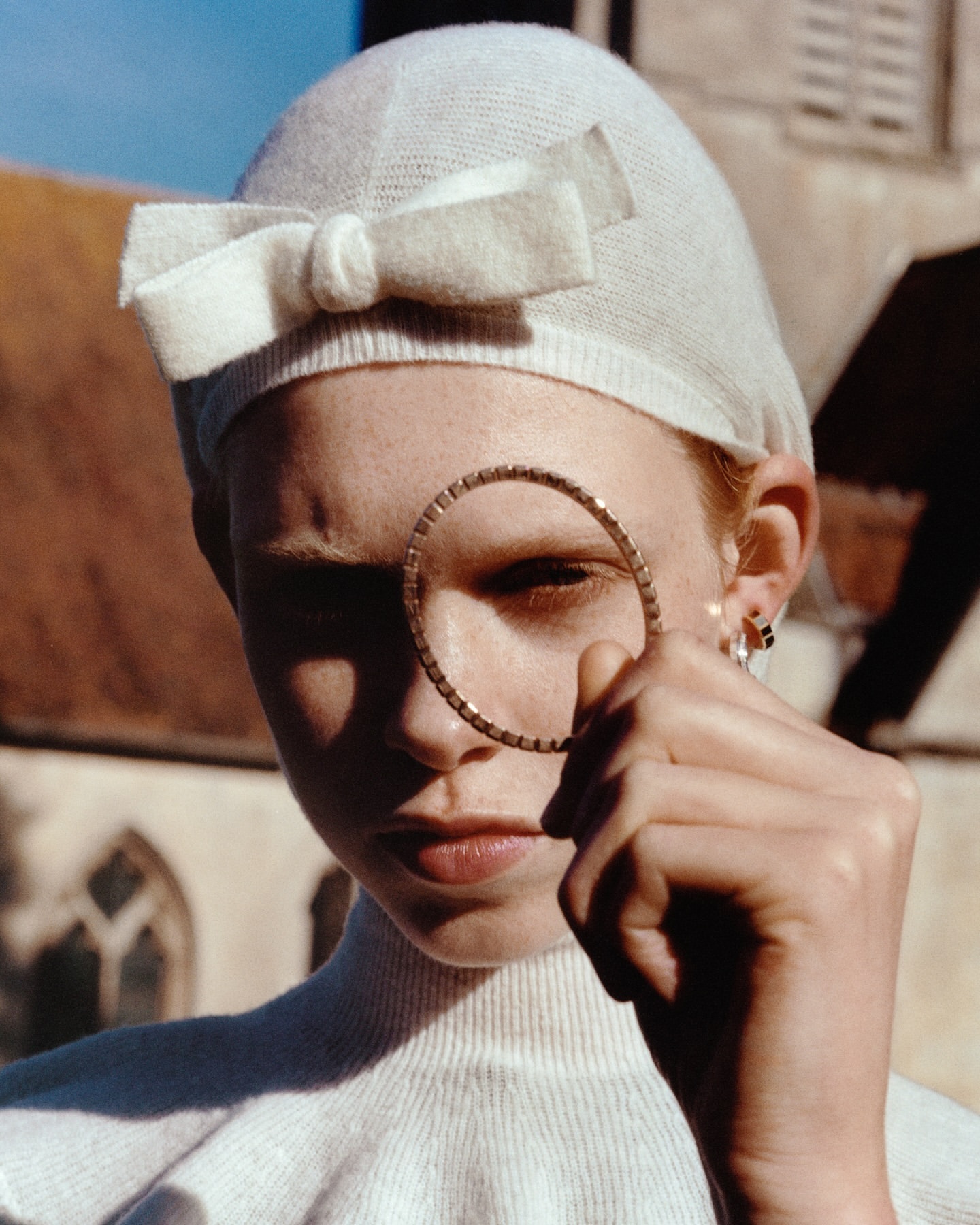 Close-up portrait of a redhead woman wearing a knit hat with a bow, holding a gold hoop earring in front of her face against a building.