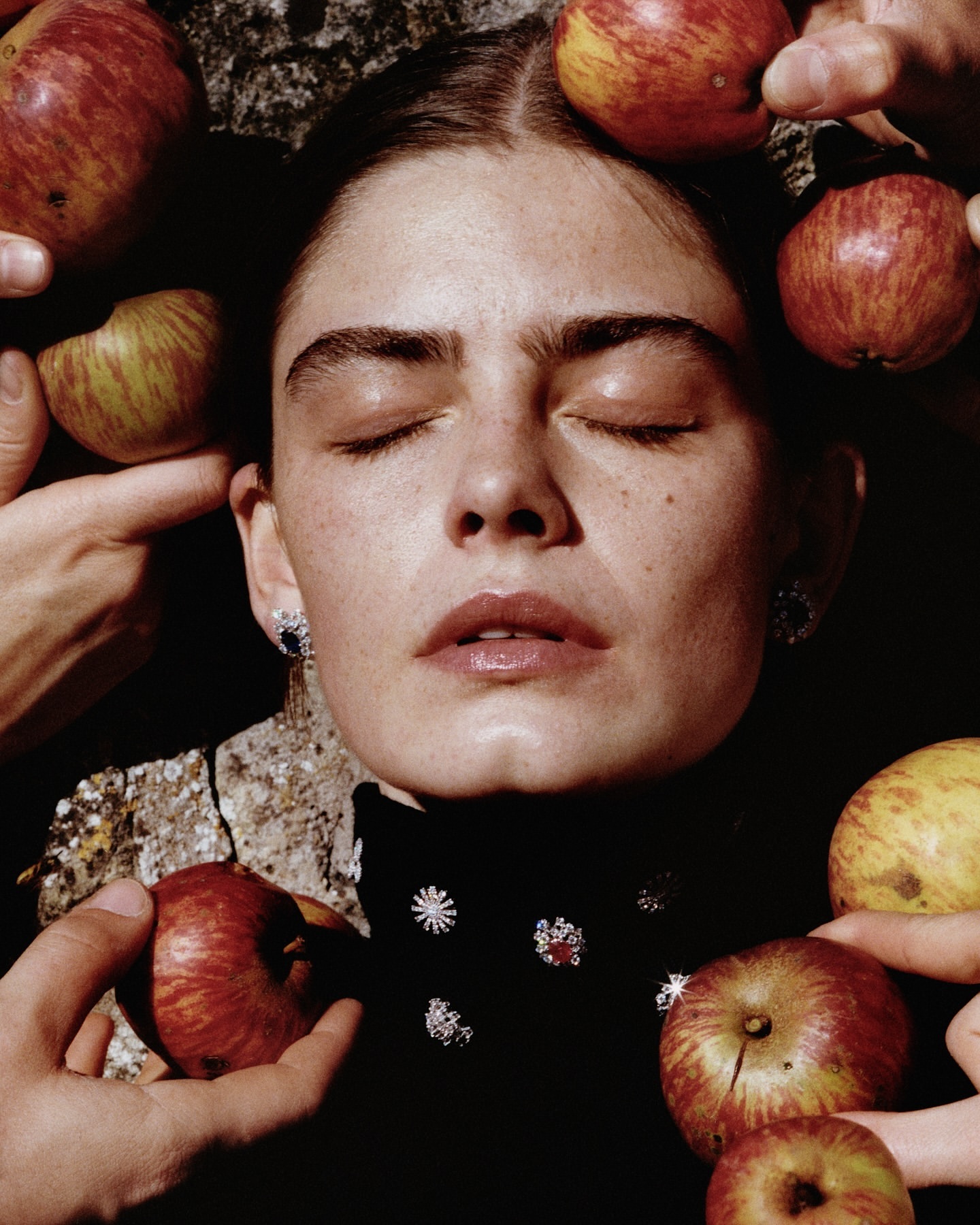 Portrait of a woman with closed eyes, surrounded by apples and hands. She has fair skin and wears a black top with embellishments.
