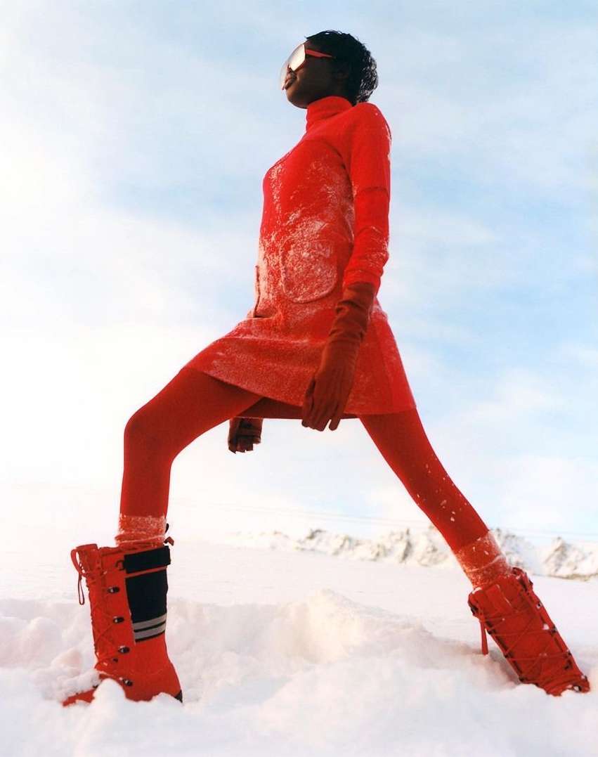 Fashionable woman in red outfit posing in snowy landscape with retro ski goggles and boots, against a cloudy sky backdrop.
