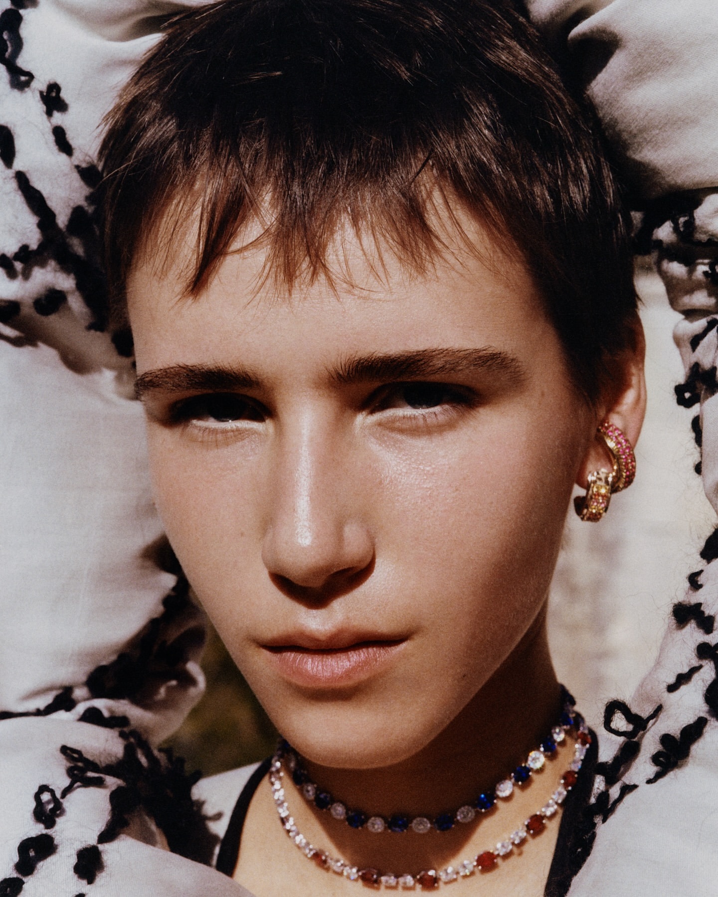 Close-up portrait of a person with short brown hair wearing layered diamond and gemstone necklaces and hoop earrings, photographed by Tom Johnson.
