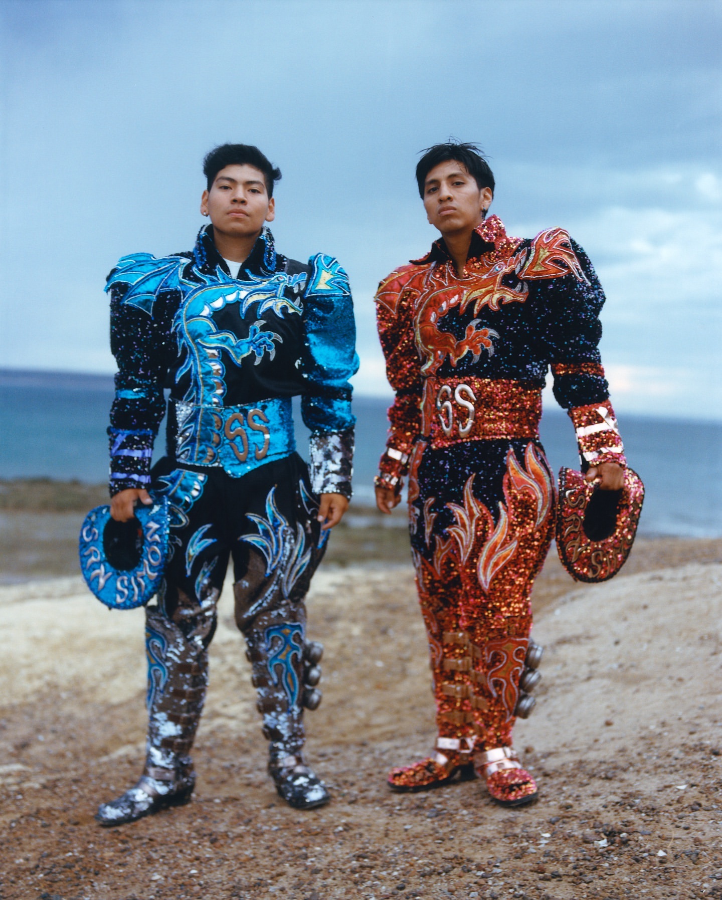 Two performers in vibrant, ornate costumes stand on a sandy shore with the ocean and sky as their backdrop. Cultural performance.