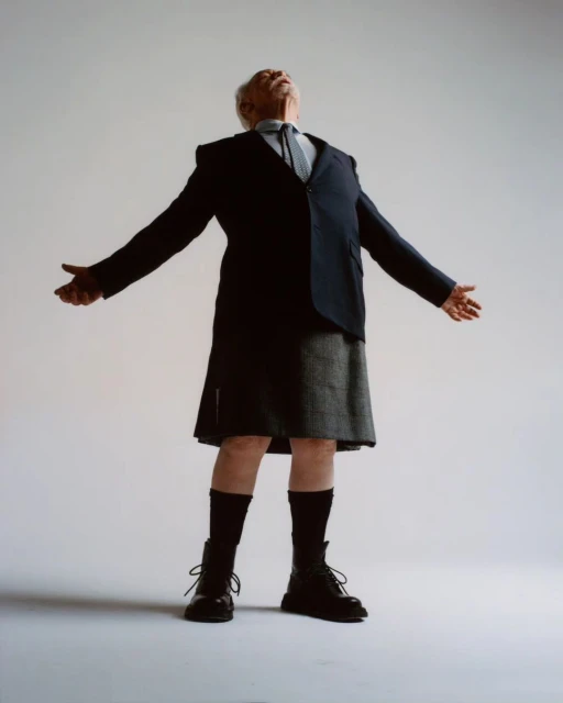 Elderly man in a suit jacket, kilt, and boots stands with arms outstretched in a studio, wearing socks and looking up.