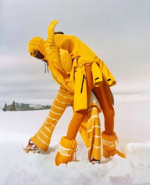 Two fashion models in bright yellow ski suits pose in snow against a cloudy sky, showcasing contemporary winter apparel.