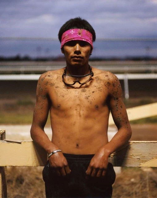 Portrait of a shirtless young man with a pink headband and goggles, standing near a wooden fence, with dirt on his body.